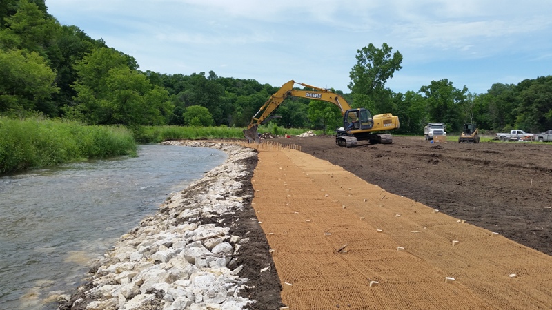 Trout stream with a track backhoe placing rock along a bank which has some bare ground and a strip of mulching fabric to prevent erosion.